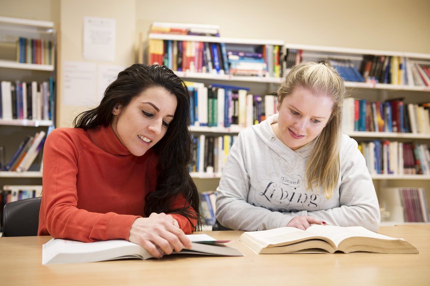 Zwei junge Frauen lernen gemeinsam in einer Bibliothek mit offenen Büchern und Bücherregalen im Hintergrund.
