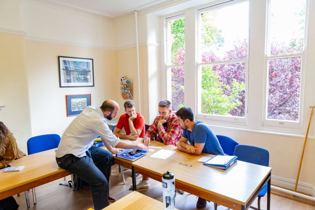 Lehrer erklärt einer Gruppe von Schülern eine Aufgabe in einem hellen Klassenzimmer mit großen Fenstern.