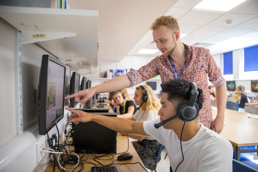 Lehrer unterstützt einen Schüler mit Headset bei einer digitalen Lernübung im Computerraum.