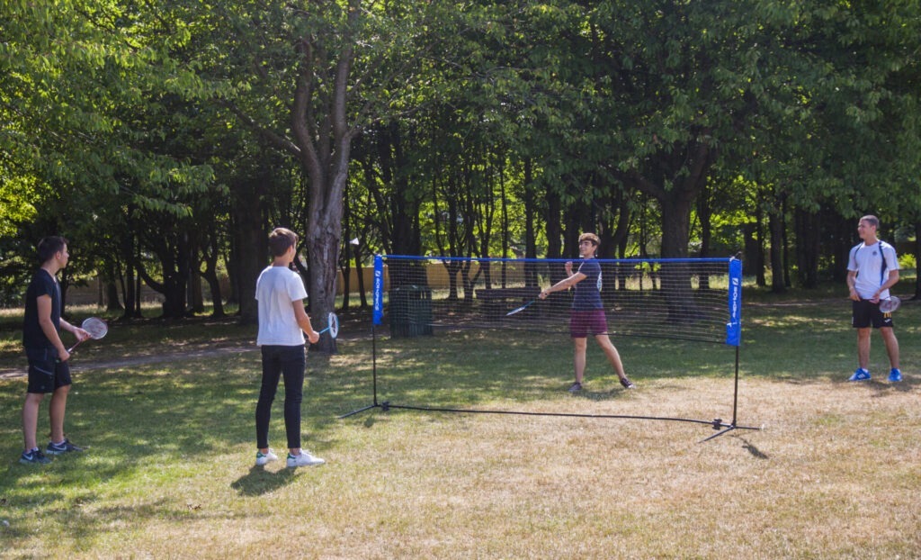 Vier junge Männer spielen Badminton auf einer Wiese in einem schattigen Park an einem sonnigen Tag.