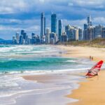 Goldener Sandstrand mit Wellen, Rettungsflaggen und einem Windsurfer, vor der Skyline einer modernen Küstenstadt.