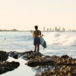 Surfer mit Surfbrett steht auf felsigem Ufer und blickt auf das Meer, mit Skyline einer Stadt im Hintergrund.