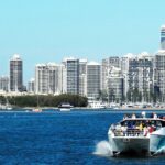 Boot mit Touristen fährt durch blaues Wasser vor der beeindruckenden Skyline einer modernen Küstenstadt.