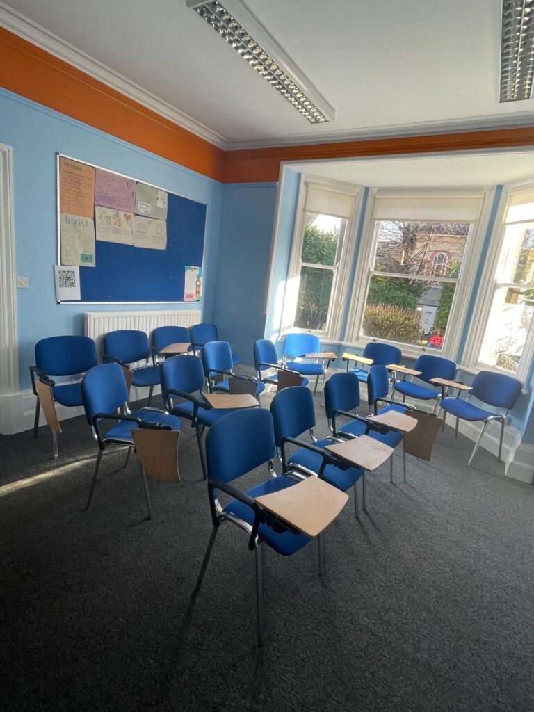Heller Klassenraum mit blauen Stühlen und Schreibbrettern, großer Fensterfront und Pinnwand.