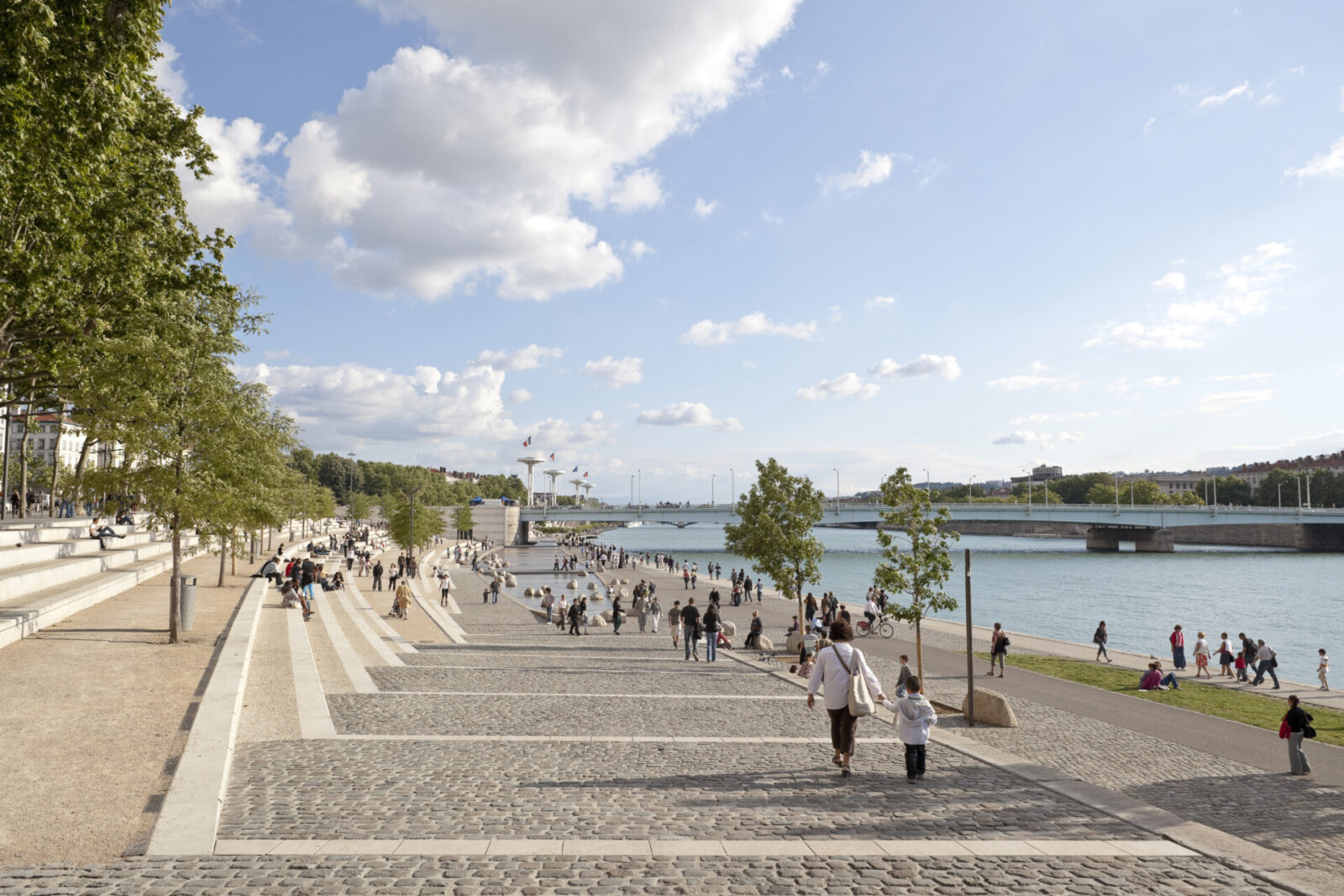 Belebte Uferpromenade mit breiten Treppen, Spaziergängern und Blick auf den Fluss sowie eine moderne Brücke.