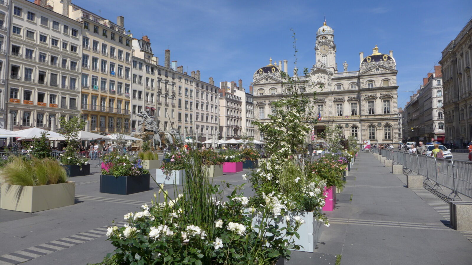 Historischer Stadtplatz mit Blumenarrangements, Cafés und beeindruckender Architektur bei sonnigem Wetter.