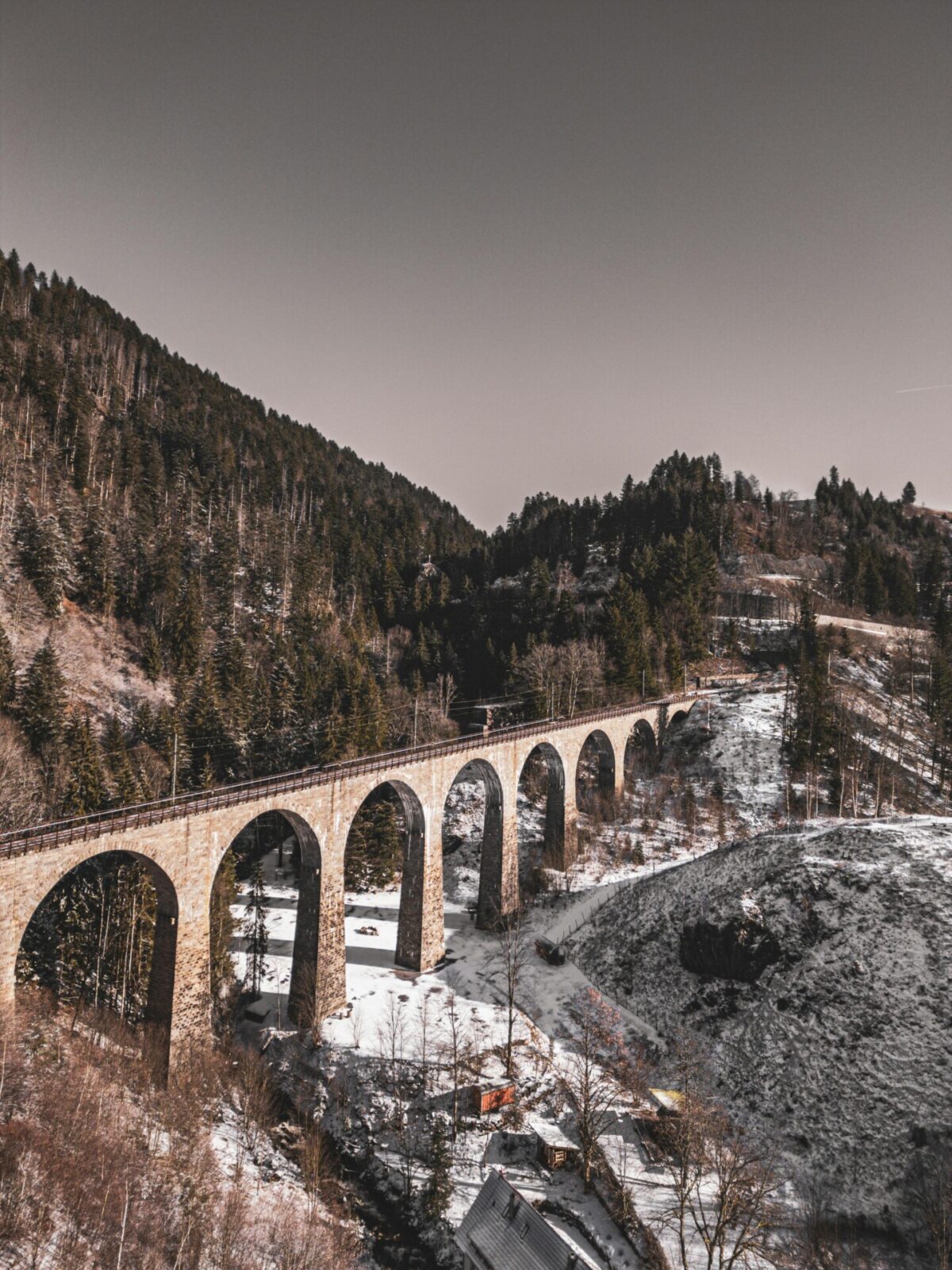 Historisches Eisenbahnviadukt in winterlicher Berglandschaft mit verschneiten Hügeln und Wäldern.