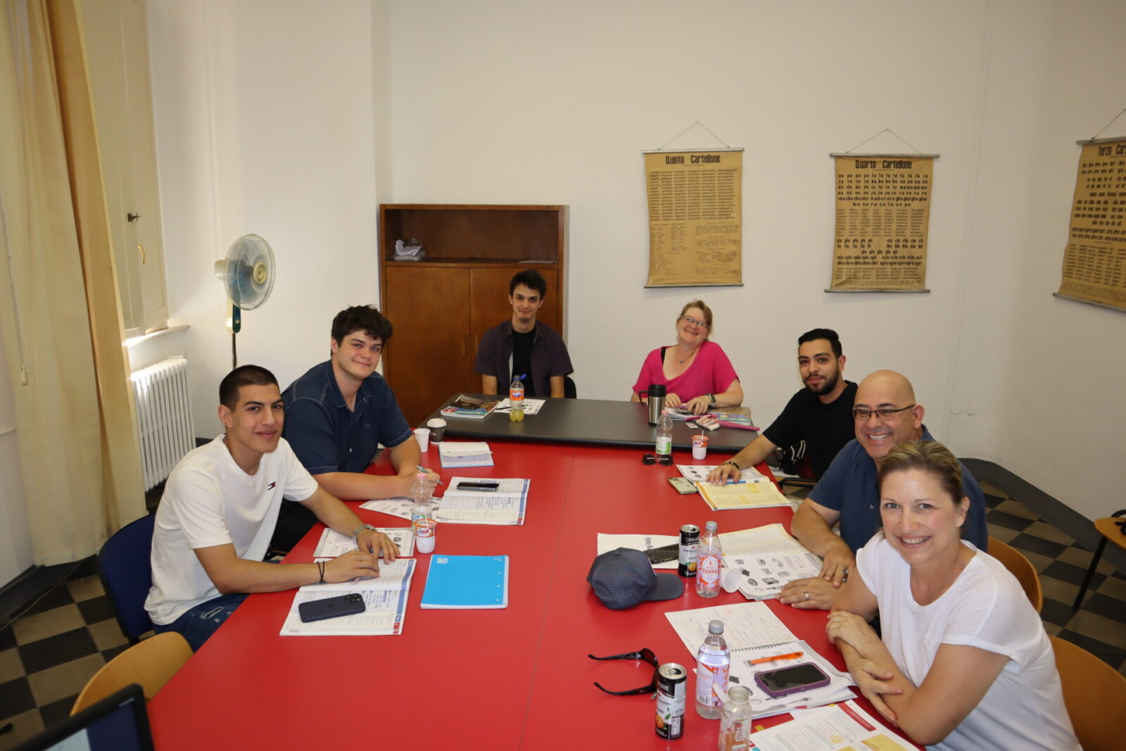 Eine Gruppe von internationalen Sprachschülern sitzt in einem Klassenzimmer an einem Tisch, lächelt und arbeitet gemeinsam an Lernmaterialien.