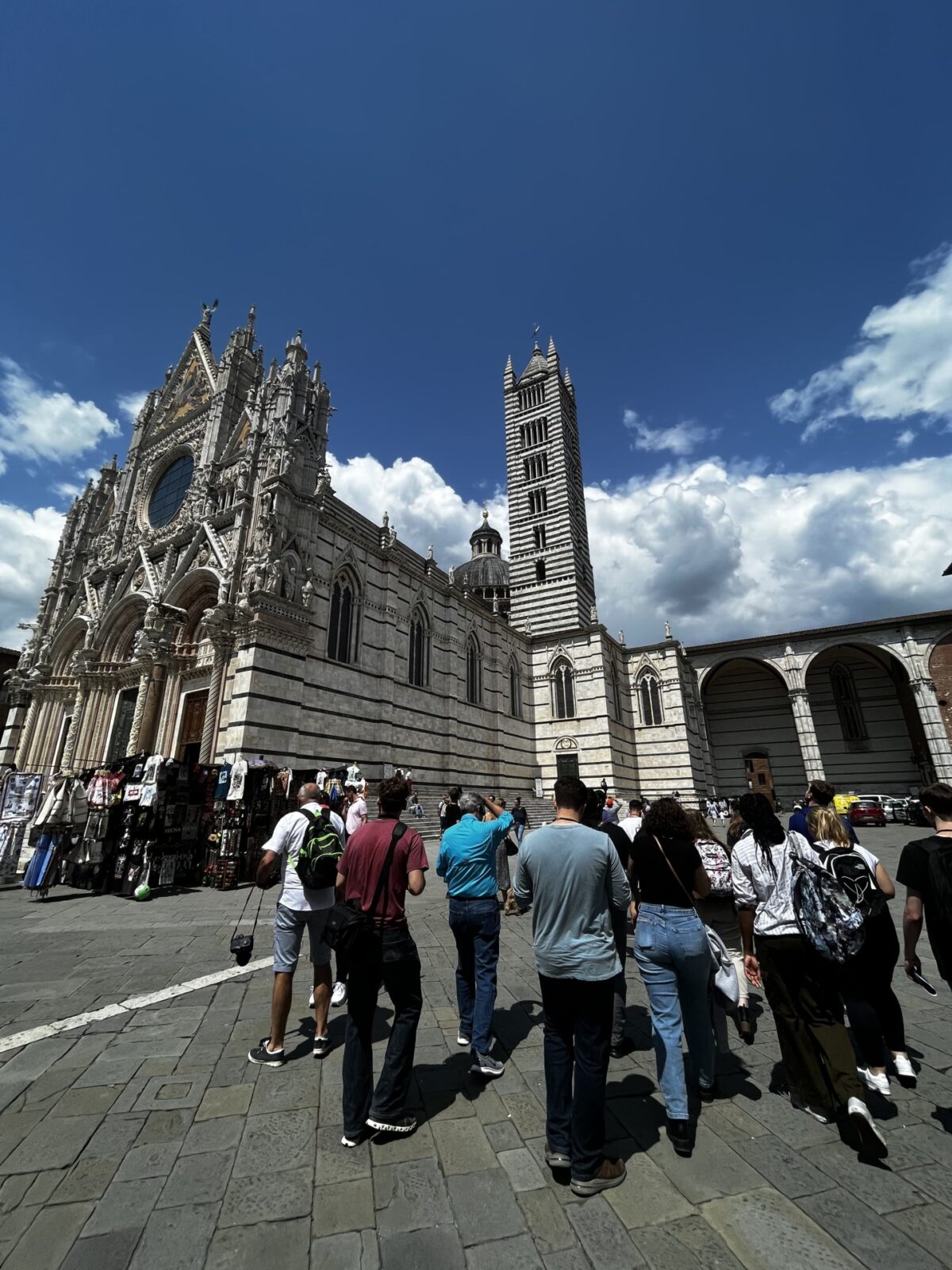 Touristen erkunden den Dom von Siena an einem sonnigen Tag mit blauem Himmel und weißen Wolken.
