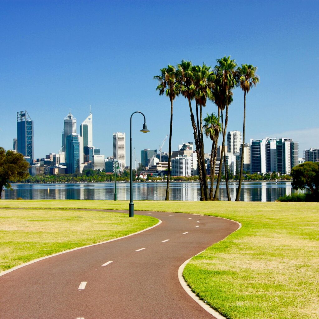 Perth Skyline mit Blick vom Park über den Swan River, umgeben von Palmen und einem Fahrradweg.