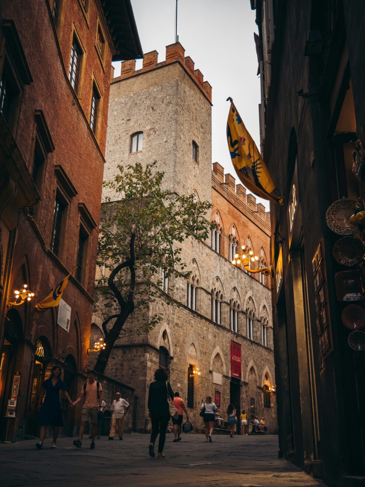 Mittelalterliche Gasse in Siena mit historischen Gebäuden, beleuchteten Laternen und wehenden Fahnen.