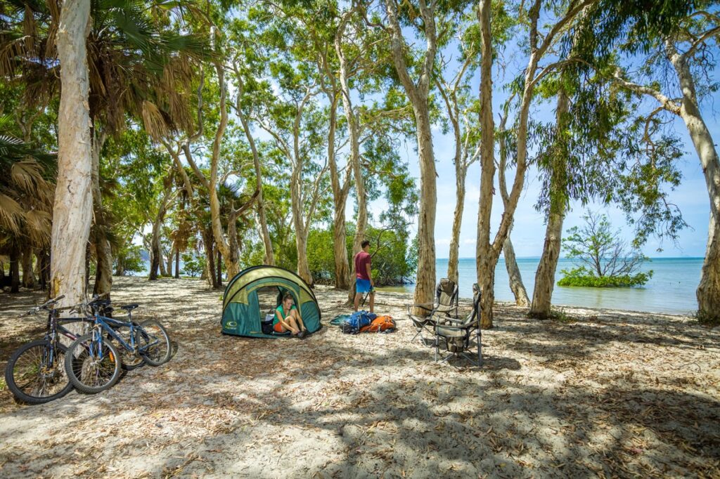 Campingplatz am tropischen Strand mit Zelt, Fahrrädern und Outdoor-Ausrüstung unter schattigen Bäumen direkt am Meer.