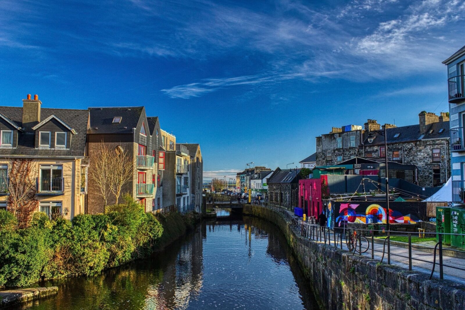 Malerischer Fluss in Galway, Irland, mit bunten Gebäuden und Spiegelungen im Wasser.