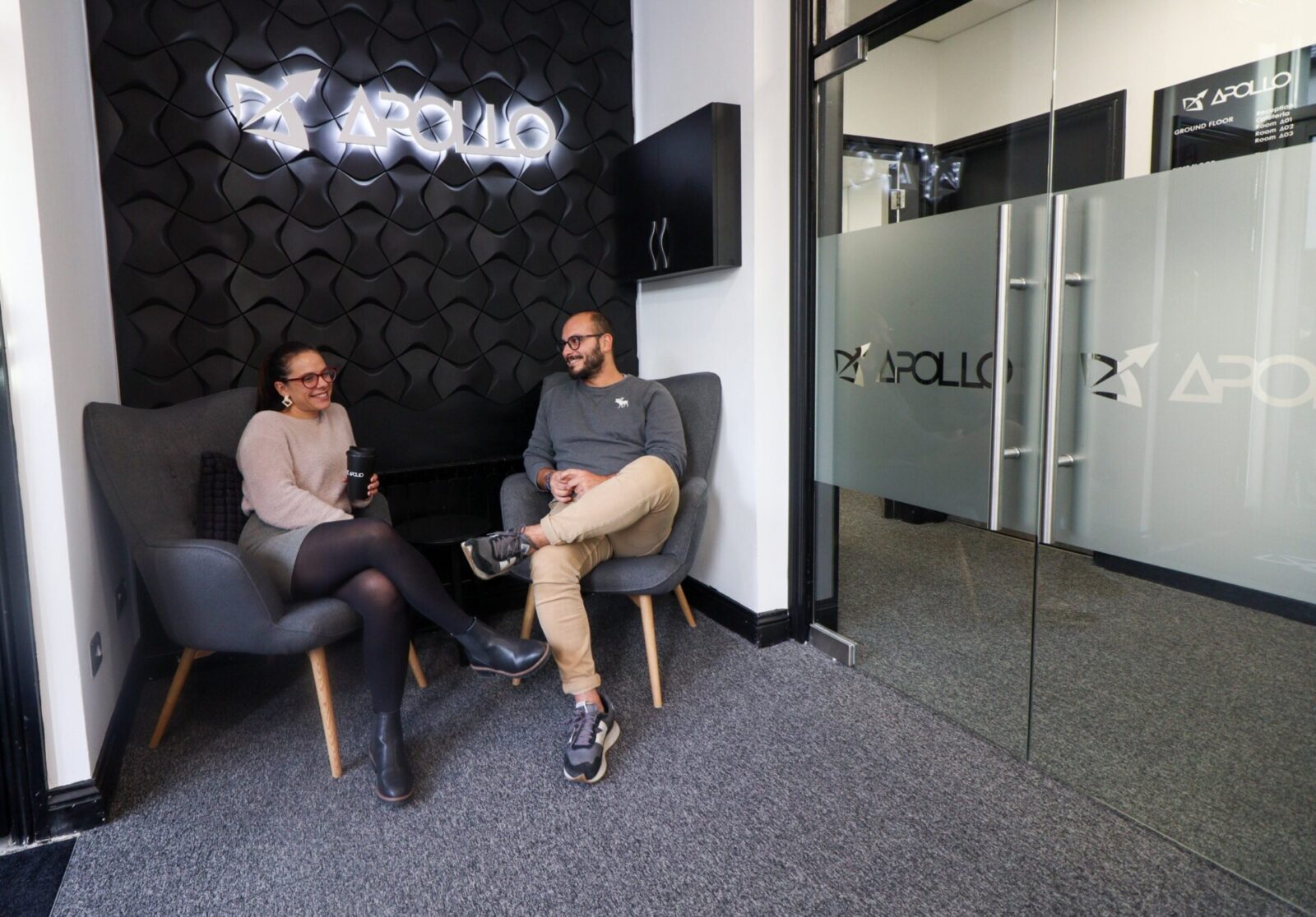 Zwei Mitarbeiter von Apollo unterhalten sich entspannt in der modernen Lounge mit stilvollem Design und beleuchtetem Firmenlogo.