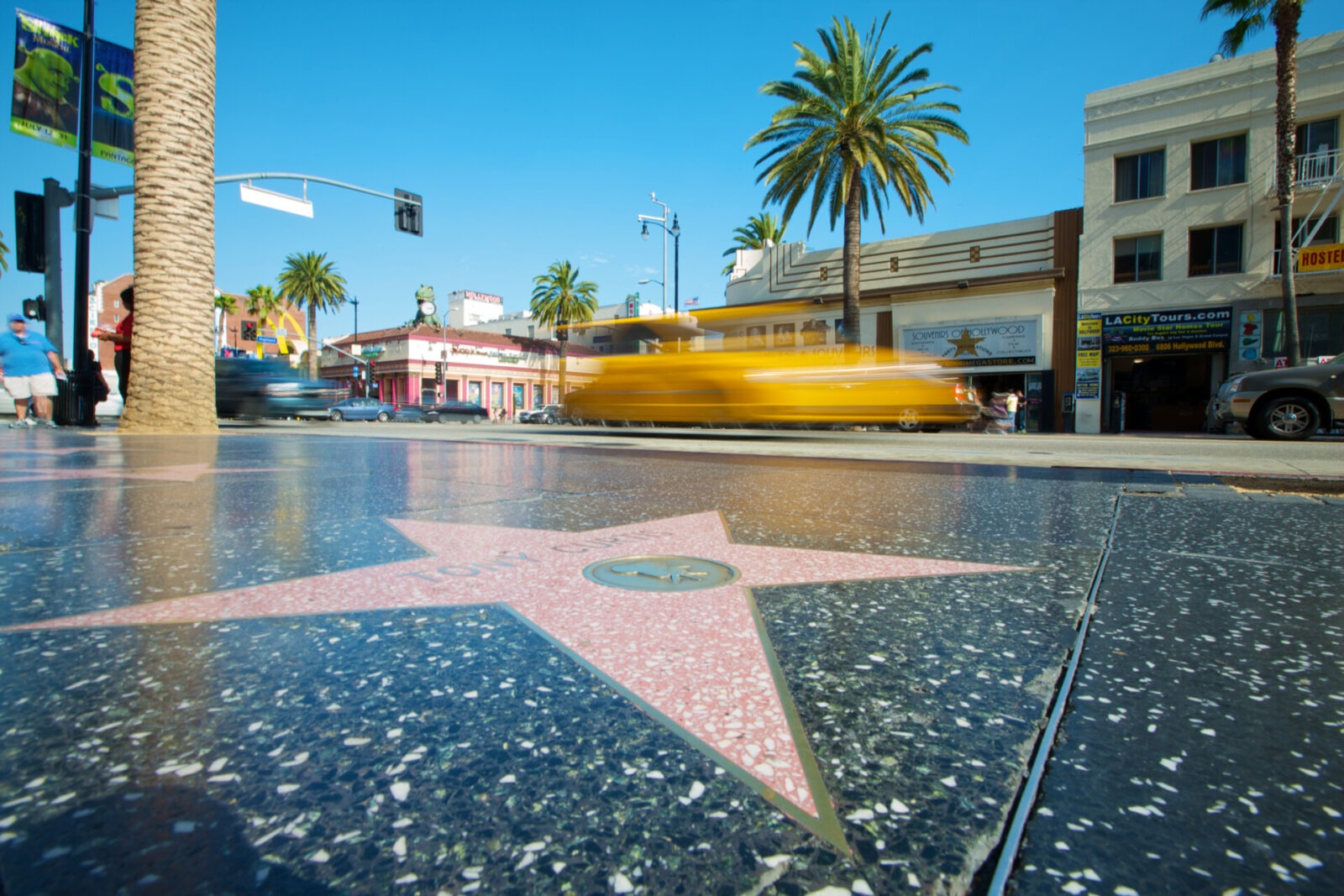 Nahaufnahme eines Sterns auf dem Walk of Fame in Hollywood mit vorbeifahrendem gelbem Taxi und Palmen im Hintergrund.
