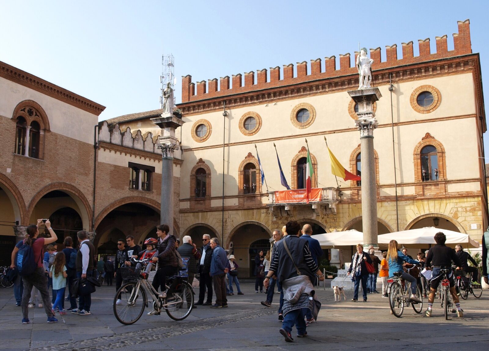 Lebendiger Platz in Italien mit historischer Architektur, Statuen und Menschen, die spazieren und Fahrrad fahren.