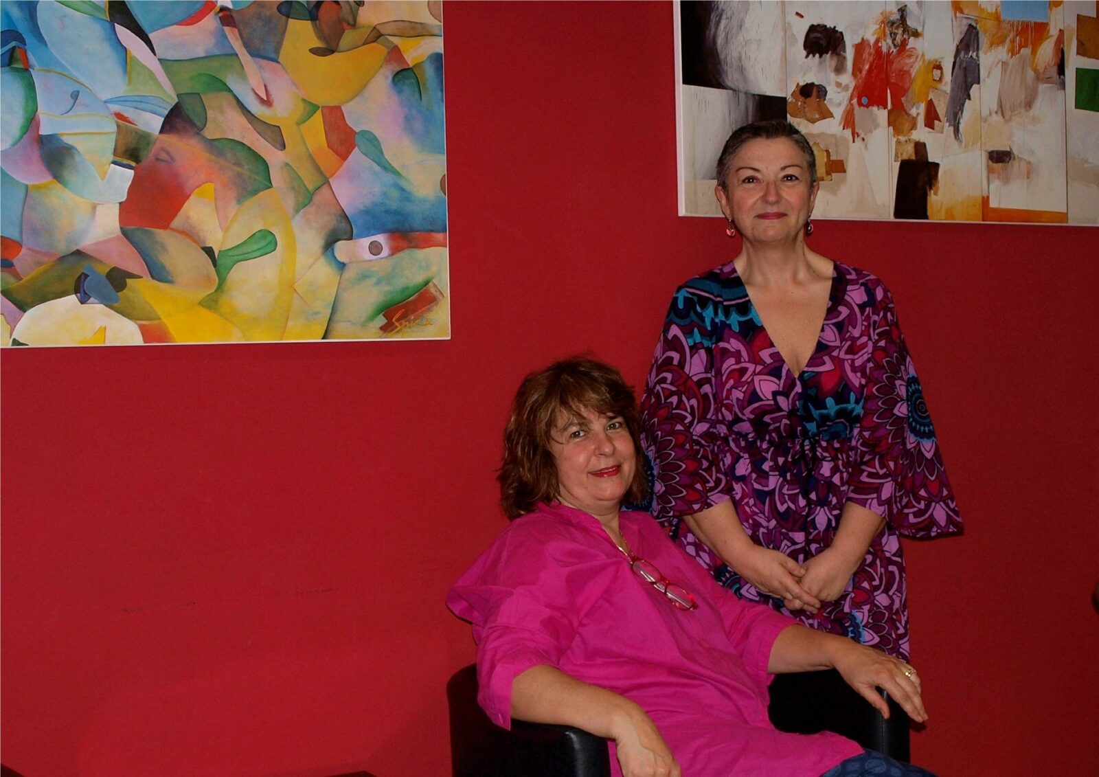 Zwei Frauen in farbenfroher Kleidung vor einer roten Wand mit moderner Kunst.